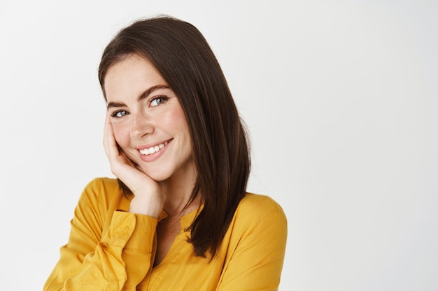 Primer plano de una linda mujer sonriente mirando coqueta a la cámara, tocando la mejilla y ruborizándose, de pie sobre la pared blanca con camisa amarilla