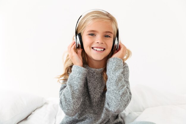 Primer plano de linda niña sonriente en auriculares sentado en la cama