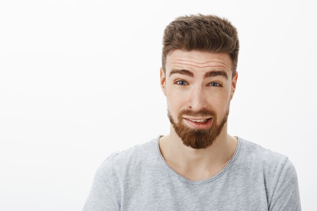 Primer plano de un lindo novio culpable con barba y peinado marrón levantando las cejas tratando de disculparse por cometer un error diciendo lo siento sintiéndome incómodo y confundido contra la pared blanca