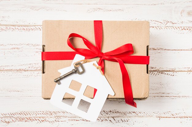Primer plano de una llave de casa atada con una cinta roja en una caja de regalo marrón sobre un escritorio de madera pintado