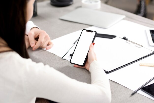 Primer plano de la mano de la empresaria sosteniendo un teléfono móvil con pantalla blanca sobre el escritorio