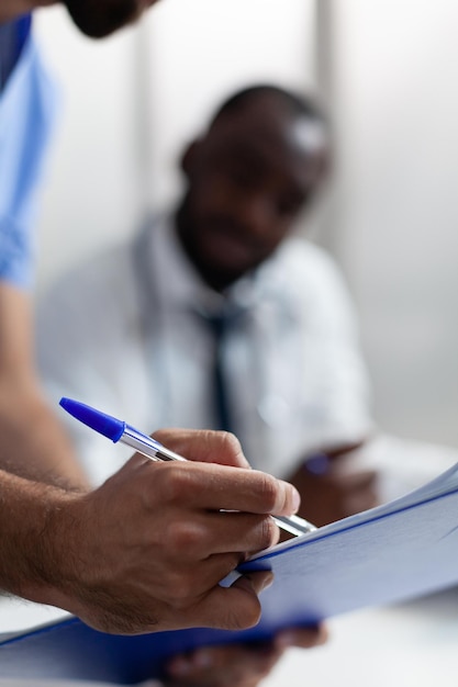 Primer plano de la mano de la enfermera del hombre que controla los síntomas de la enfermedad en el portapapeles discutiendo el tratamiento médico con el médico que trabaja en la oficina del hospital. Médico afroamericano analizando documentos médicos