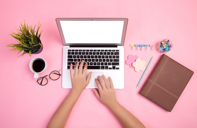 Primer plano de la mano escribiendo en la computadora portátil con papelería y una taza de café sobre fondo rosa