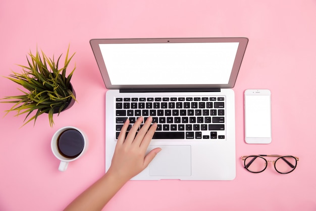 Primer plano de la mano escribiendo en la computadora portátil con papelería y una taza de café sobre fondo rosa