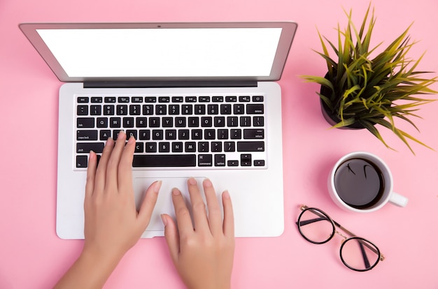 Primer plano de la mano escribiendo en la computadora portátil con papelería y una taza de café sobre fondo rosa