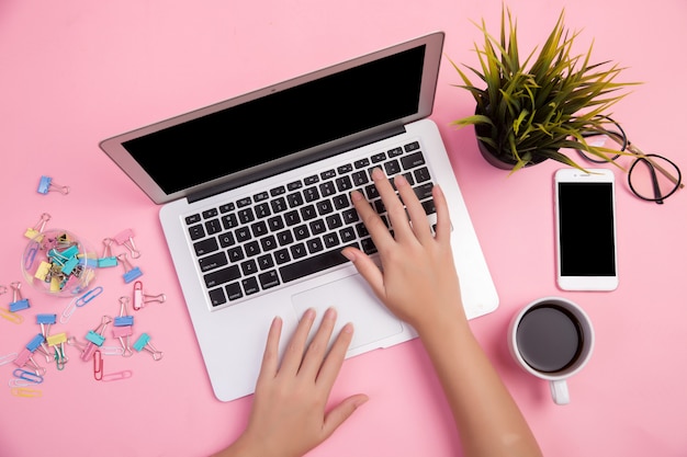 Primer plano de la mano escribiendo en la computadora portátil con papelería y una taza de café sobre fondo rosa