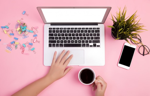 Primer plano de la mano escribiendo en la computadora portátil con papelería y una taza de café sobre fondo rosa