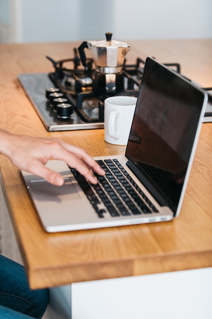 Primer plano de la mano escribiendo en la computadora portátil sobre el mostrador de la cocina de madera con taza de café