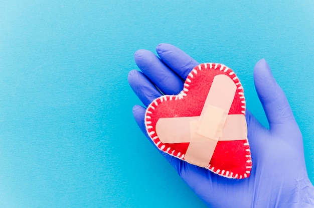 Primer plano de la mano con guantes quirúrgicos que sostienen el corazón cosido en forma de corazón de la materia textil con los vendajes cruzados en el contexto azul