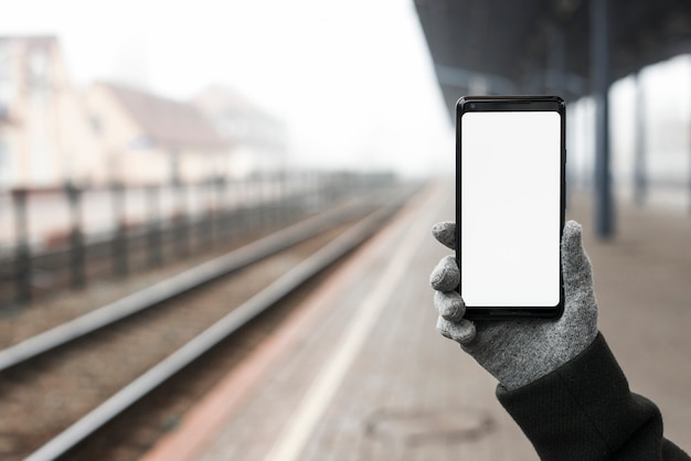 Primer plano de la mano con guantes sosteniendo un teléfono móvil que muestra una pantalla blanca en blanco en la estación de tren