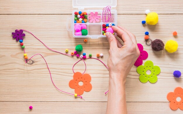 Primer plano de mano haciendo collar con perlas; bola de flores y algodón en el fondo con textura de madera