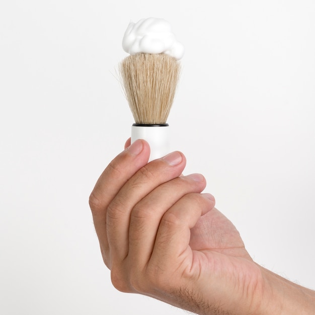 Primer plano de la mano del hombre con brocha de afeitar y espuma contra la pared blanca