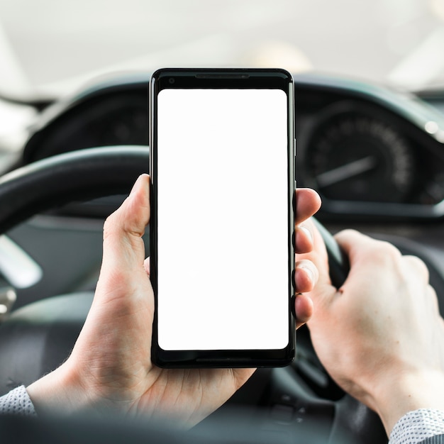 Primer plano de la mano del hombre conduciendo un automóvil que muestra un teléfono móvil con pantalla blanca en blanco