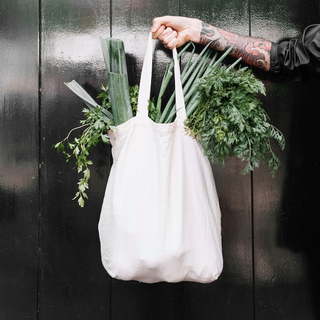 Primer plano de la mano del hombre que sostiene la bolsa de la compra blanca llena de verduras de hoja