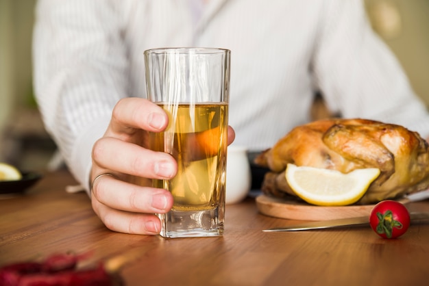 Primer plano de la mano de un hombre que sostiene el vaso de una cerveza con pollo asado entero en la mesa