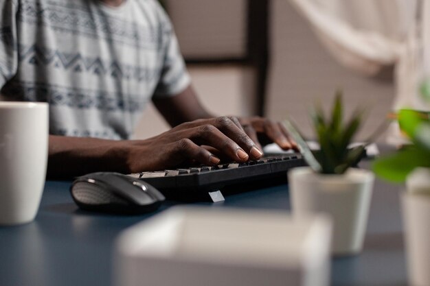 Primer plano de la mano del joven empleado afroamericano navegar por la información de gestión escribiendo ideas de proyectos en internet usando la computadora sentado en el escritorio en la sala de estar. Emprendedor que trabaja lejos de casa