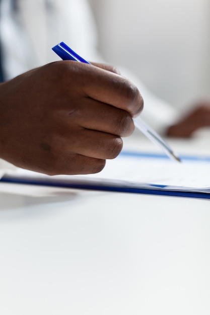 Primer plano de la mano de un médico afroamericano que analiza los documentos de prescripción de medicamentos que trabajan en el tratamiento de la salud en el consultorio del hospital. Hombre terapeuta monitoreando los síntomas de enfermedad del paciente