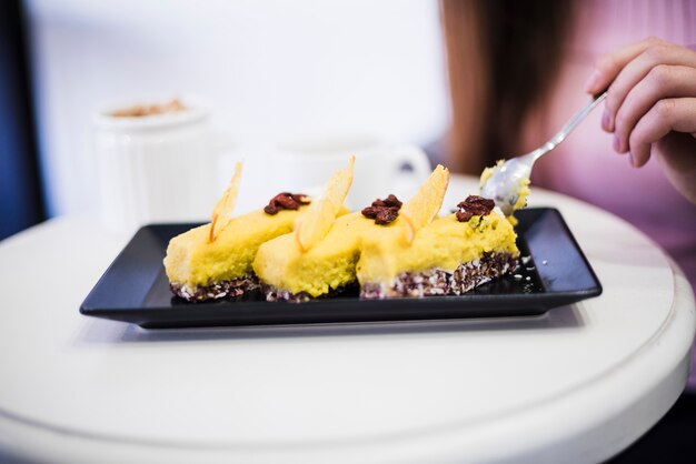 Primer plano de la mano de una mujer comiendo rebanada de pastel con una cuchara en una bandeja negra sobre la mesa blanca