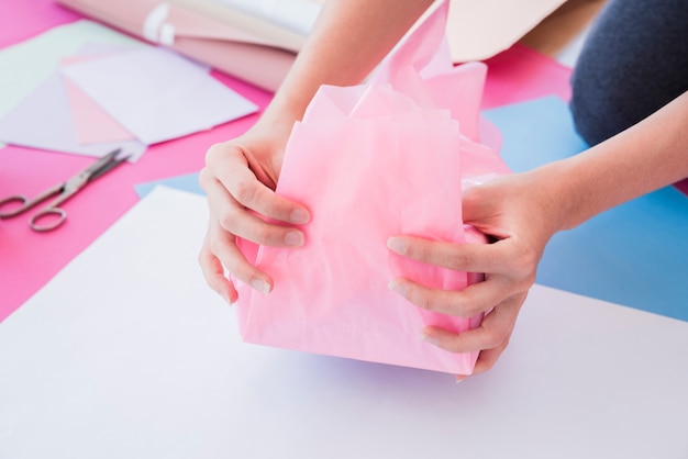 Primer plano de la mano de una mujer envolviendo papel rosa en una caja de regalo