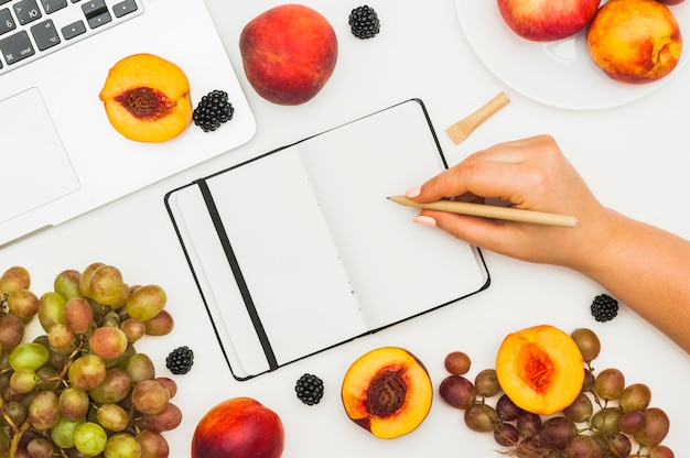 Primer plano de la mano de una mujer escribiendo en un diario con frutas y una computadora portátil en la mesa blanca