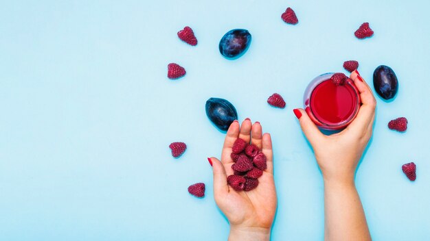 Primer plano de la mano de la mujer con frambuesas y jugo con ciruelas sobre fondo azul