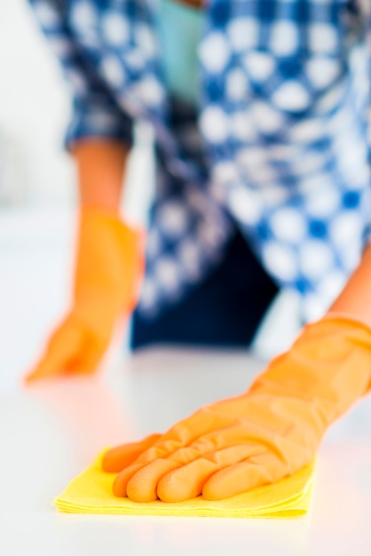 El primer plano de la mano de la mujer con guantes amarillos limpia la superficie blanca con una servilleta amarilla