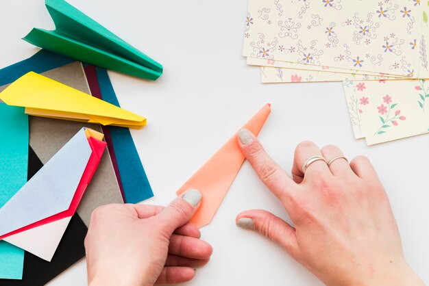 Primer plano de la mano de la mujer haciendo un avión de papel con papeles de colores en el escritorio blanco