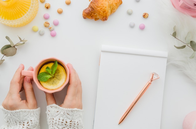 Foto gratuita primer plano de la mano de una mujer que bebe el té de limón con la pluma y la libreta en blanco en la tabla blanca