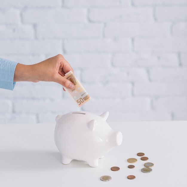 Primer plano de la mano de una mujer que inserta billetes de cincuenta euros en piggybank en el escritorio