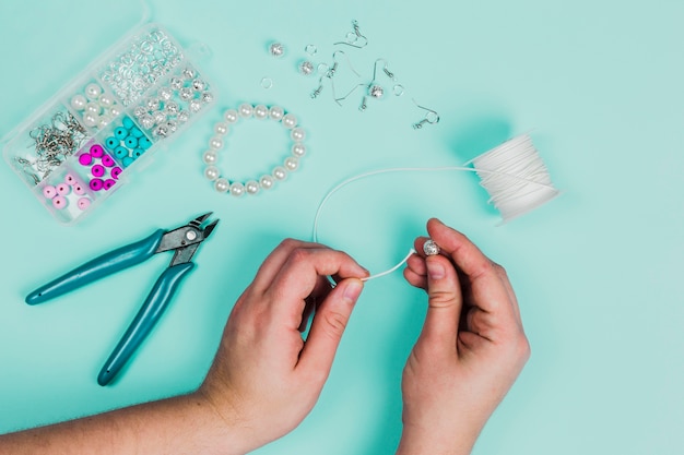 Primer plano de la mano de la mujer que inserta la perla en el hilo blanco para hacer la pulsera sobre fondo verde azulado