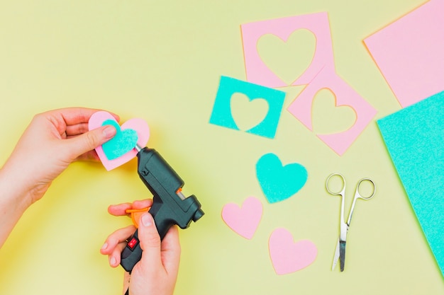 Primer plano de la mano de la mujer que pega la forma de corazón de papel con una pistola eléctrica de pegamento caliente