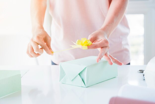 Primer plano de la mano de la mujer que pega el lazo amarillo en la caja de regalo envuelta sobre la mesa