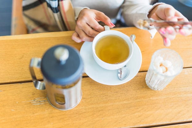 Primer plano de la mano de una mujer que pone azúcar moreno con las pinzas en el té de hierbas en la mesa de madera