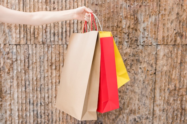 Primer plano de la mano de una mujer que sostiene bolsos de compras multicolores delante de la pared marrón