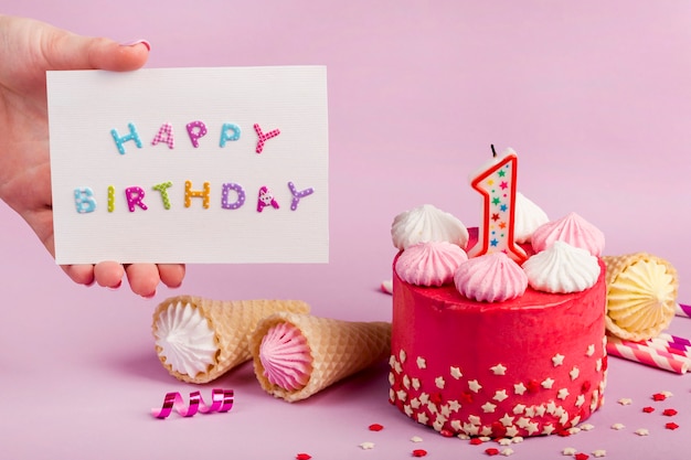 Primer plano de la mano de una mujer que sostiene la tarjeta de feliz cumpleaños cerca de la torta decorativa contra el telón de fondo púrpura