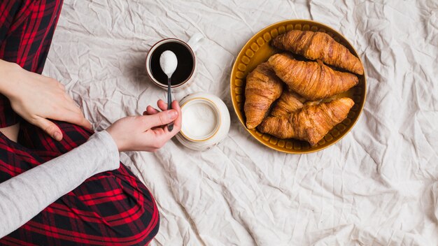 Primer plano de la mano de la mujer que vierte la leche en polvo en la taza de té con croissant horneado en la sábana desmenuzada