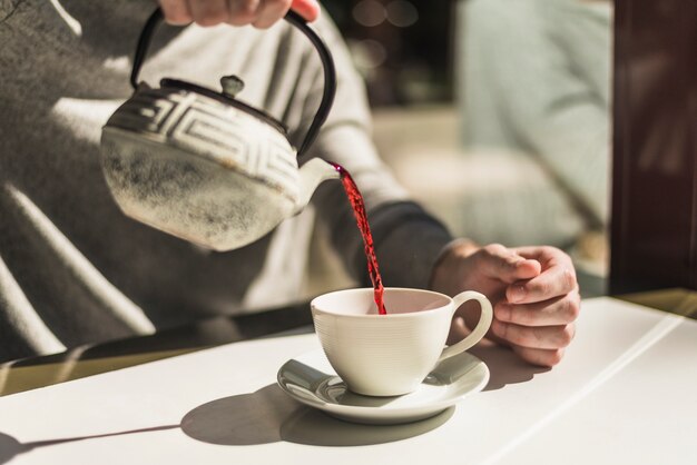 Primer plano de la mano de una mujer que vierte té rojo de caldera tradicional en la taza blanca