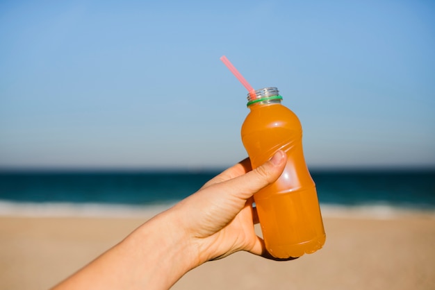 Primer plano de la mano de la mujer sosteniendo una botella de plástico de jugo de naranja con pajita en la playa