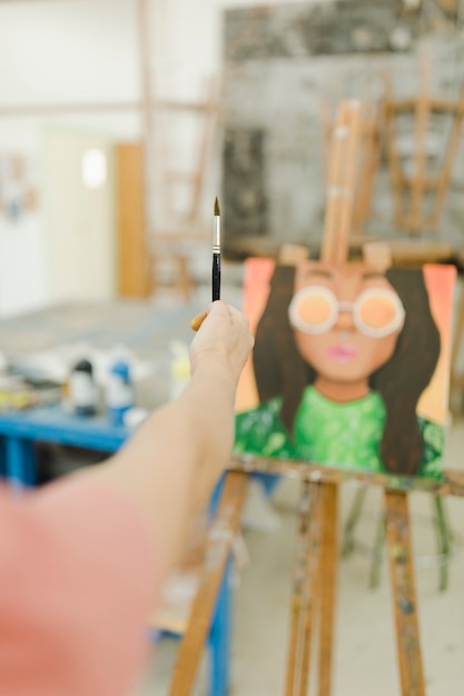 Primer plano de la mano de mujer sosteniendo el pincel delante de la pintura en el caballete