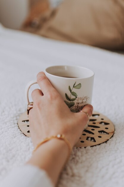 Primer plano de la mano de una mujer sosteniendo una taza blanca con una pintura puesta sobre una superficie blanca