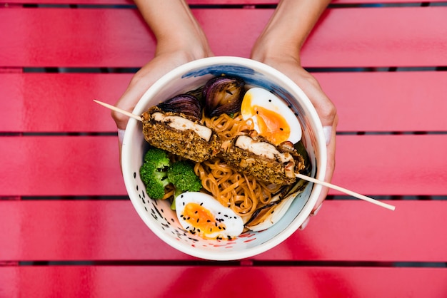 Primer plano de la mano de una mujer sosteniendo un tazón de fideos ramen con huevo; cebolla y brócoli en rojo