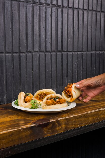 Primer plano de la mano de la mujer tomando gua bao en un plato blanco sobre la mesa de madera