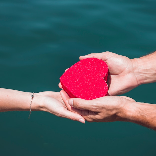 Primer plano de la mano de una pareja con forma de corazón rojo frente al mar