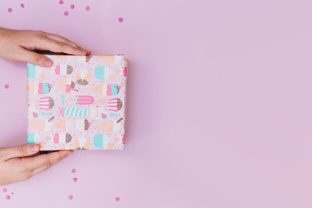 Primer plano de la mano de una persona con caja de regalo envuelta con confeti sobre fondo rosa