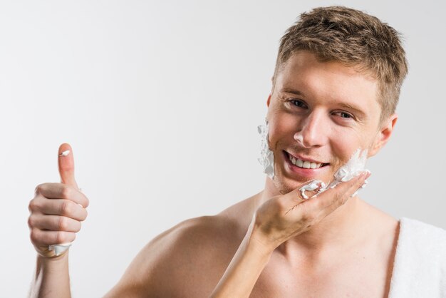 Primer plano de la mano de una persona que aplica espuma de afeitar en su mejilla mostrando el pulgar hacia arriba de la señal contra el fondo gris