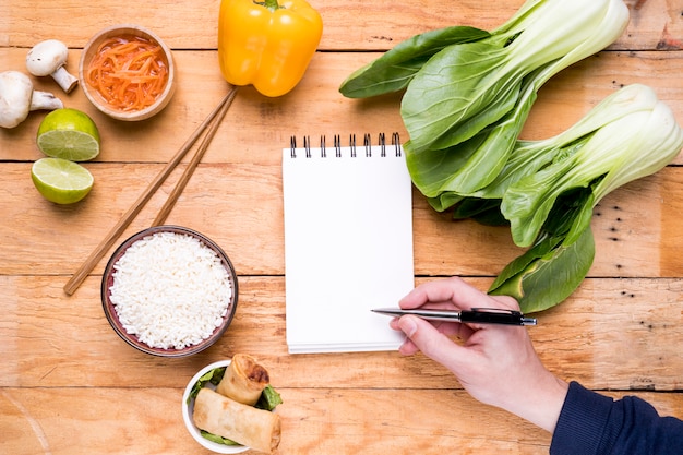 Primer plano de la mano de una persona que escribe en la libreta espiral blanca en blanco con la comida tailandesa en la mesa de madera