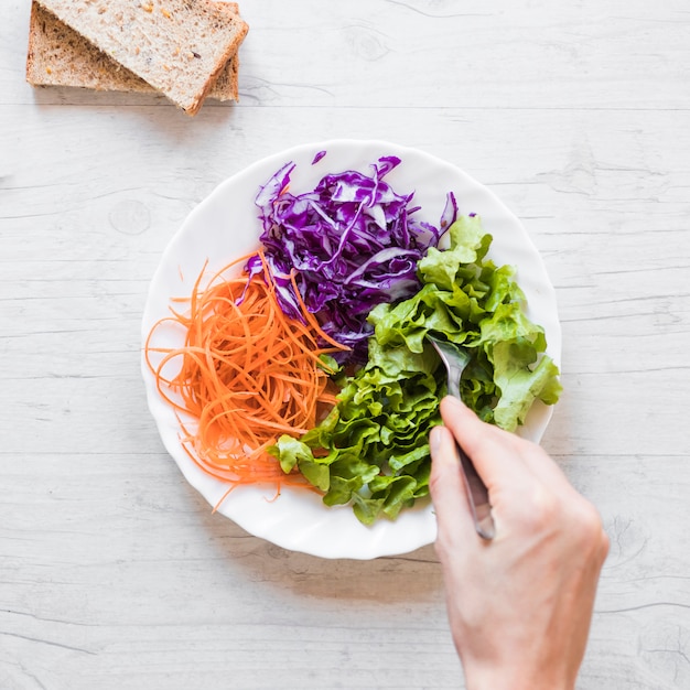 Foto gratuita primer plano de la mano de la persona que toma ensalada de verduras con una cuchara en el escritorio de madera