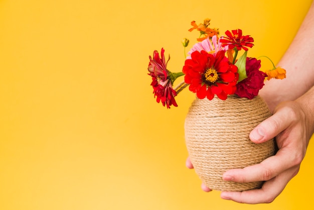 Primer plano de la mano de una persona sosteniendo un jarrón de flores sobre fondo amarillo