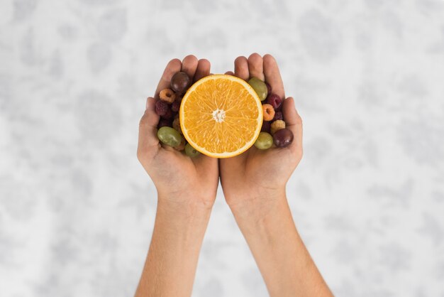 Primer plano de la mano de una persona sosteniendo una naranja a la mitad con uvas y frambuesa