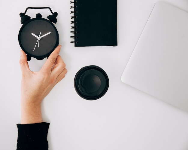 Primer plano de la mano de una persona sosteniendo un reloj despertador negro; bloc de notas espiral Taza de café portátil y para llevar en el escritorio blanco